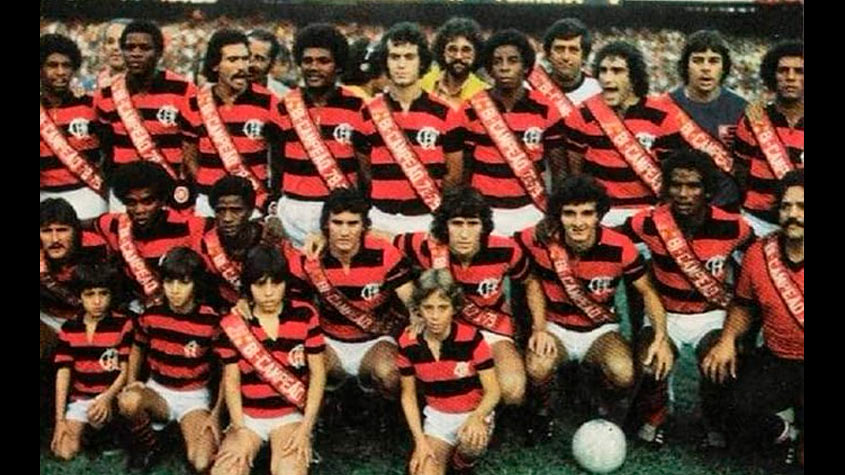 Fotografia real da comemoração do título carioca de 1979 no gramado do Maracanã, com Zico erguendo o troféu cercado por fotógrafos de campo e companheiros. Em 1979, o Flamengo de Cláudio Coutinho realizou a façanha inédita de vencer dois Campeonatos Cariocas distintos no mesmo ano civil. (Fonte Oficial: Diário Lance! & Jornal dos Sports (Acervo Fotográfico) · Fotografia de Campo Original (1979) / Acervo CRF & Lance!)